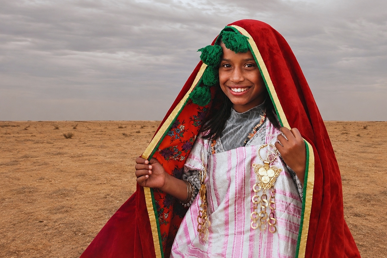 Jeune fille amazighe en costume traditionnel, souriant dans le désert près de Douz, sous un large voile rouge bordé de vert et de jaune.