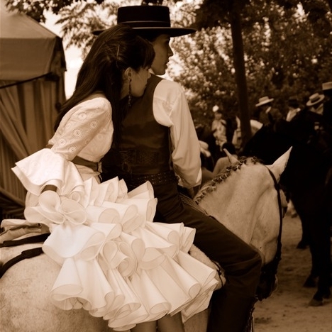 Jeune fille en amazone sur un cheval pendant la feria d’Arcos de la Frontera, portant une robe andalouse blanche à volants derrière un homme en costume.