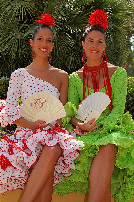 Deux jeunes femmes en robes andalouses colorées, assises devant des palmiers et tenant des éventails à la Feria de Jerez de la Frontera.