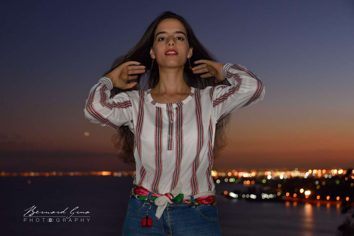 Abir portant une chemise traditionnelle tunisienne blanche à rayures rouges, devant le golfe de Tunis au coucher du soleil.