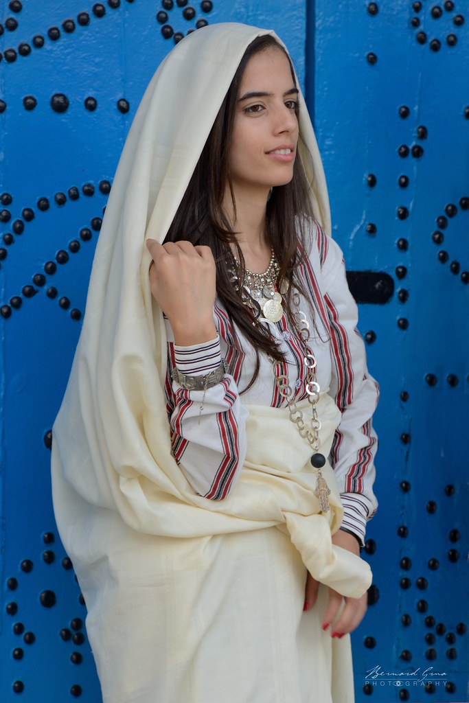Abir portant un sefsari blanc et une tenue traditionnelle tunisienne, devant une porte bleue de Sidi Bou Saïd.