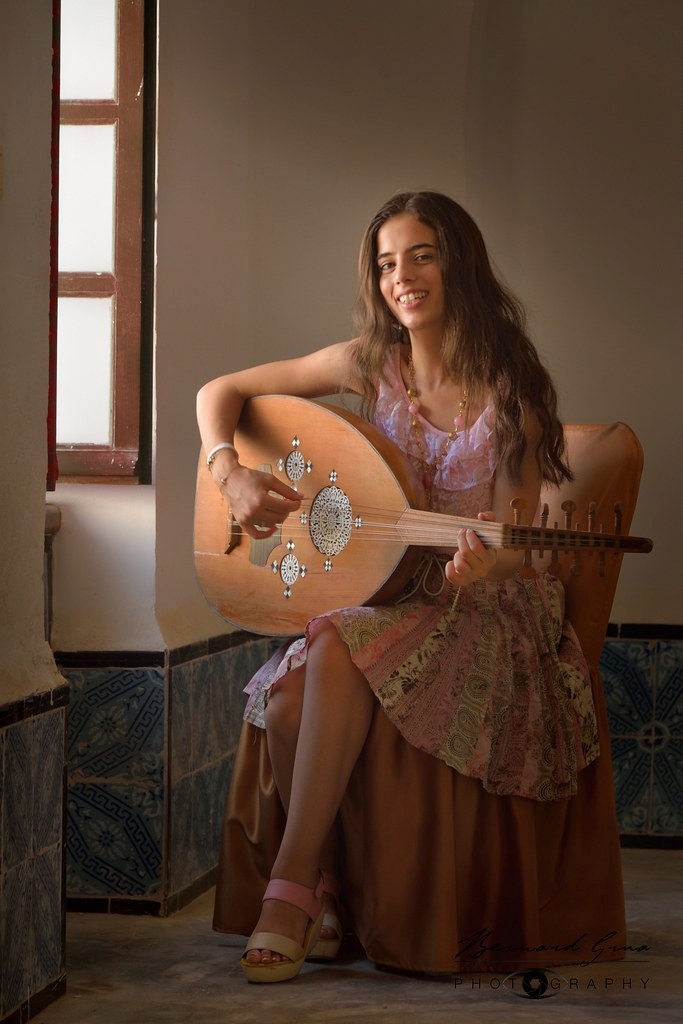 Abir assise dans un intérieur traditionnel à Mahdia, jouant du oud vêtue d'une robe rose et dorée.