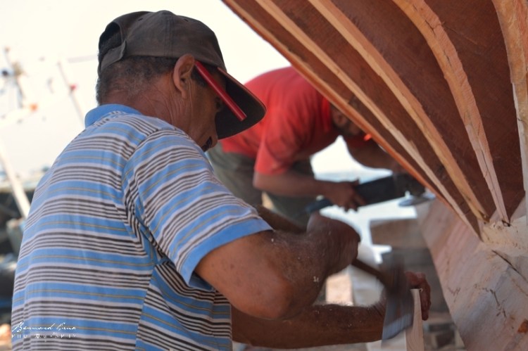 Bernard Grua - M. Fathi Arous travaille, ici avec, semble-t-il, une herminette, outil traditionnel de charpenterie de marine. El Attaya, Kerkennah, Tunisie, Chantier Naval Fathi Arous. 2 octobre 2019