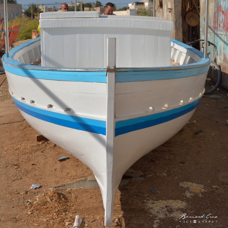 Bernard Grua - Flouka neuve terminée. Les formes sont plus fortes. On voit que ce bateau ne naviguera pas à la voile. Le fardage devient moins une préoccupation. Le pavois est plus élevé que sur les purs voiliers. Un roof (généralement amovible) couvre l'hiloire principal. Depuis quelques années, une lame d'inox arrondie couvre la bordure du plat bord. Une structure verticale en inox monte du plat bord. Chantier Naval Zouhir Chelly - El Attaya, Kerkennah, Tunisie 2 octobre 2019